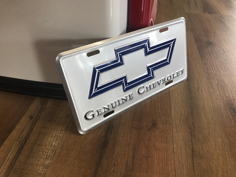 May include: A white metal license plate with a blue Chevrolet logo and the text "Genuine Chevrolet".