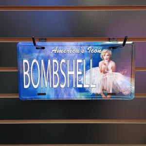 May include: A blue and white metal license plate with the text "America's Icon BOMBSHELL" and a picture of Marilyn Monroe.