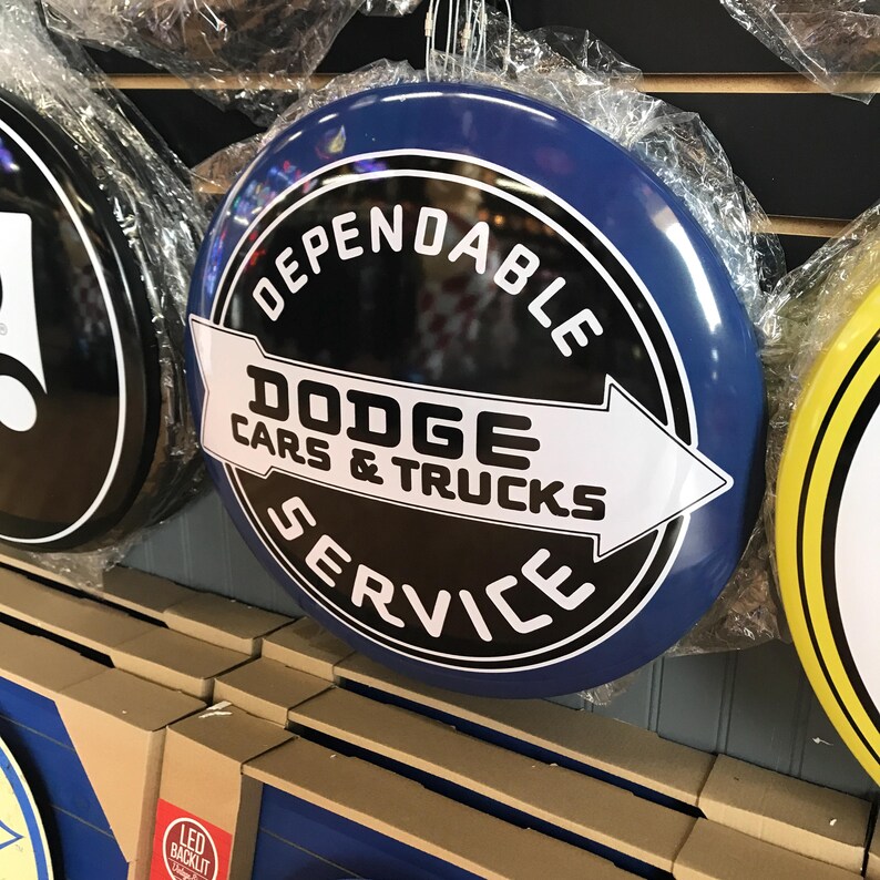 Puede incluir: Un letrero redondo de metal azul y negro con texto blanco que dice "Dependable Dodge Cars & Trucks Service". El letrero tiene una flecha blanca apuntando hacia la derecha.