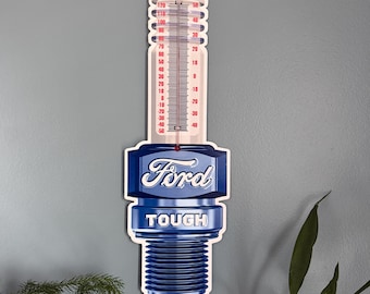 Könnte beinhalten: Ein blaues und weißes Metallschild in Form einer Zündkerze mit einem Thermometer an der Seite. Das Schild trägt die Aufschrift "Ford Tough".