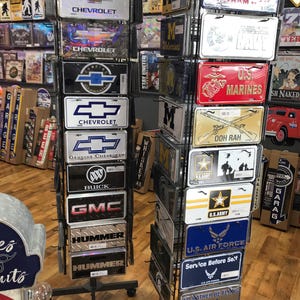 May include: A metal display rack with two rows of license plate-style signs. The top row features signs with military branch logos and text, including "Veteran", "US Marines", and "Ooh Rah". The bottom row features signs with military branch logos and text, including "U.S. Army", "U.S. Air Force", and "Service Before Self".