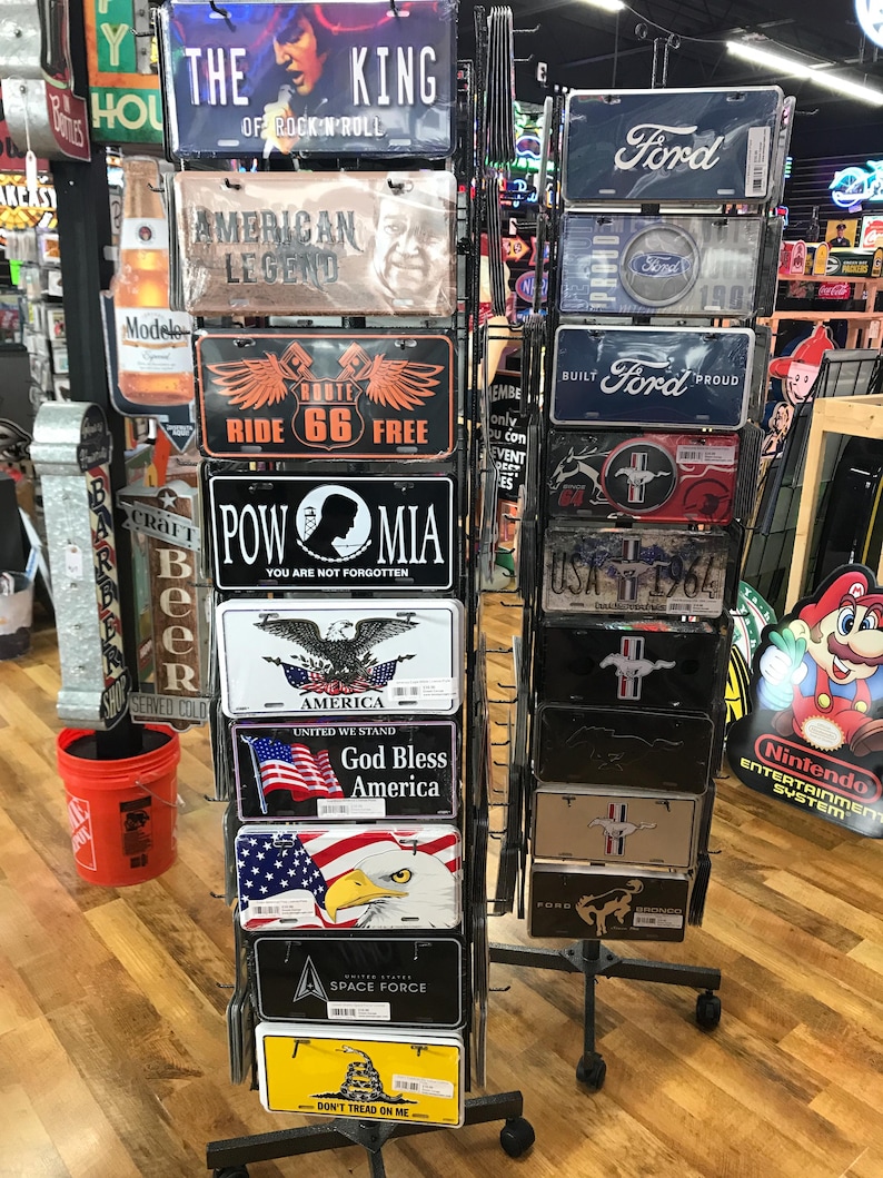 May include: A metal display rack with various metal signs. The signs feature different themes, including American patriotism, music, and automobiles. Some of the signs include text such as "The King of Rock 'n' Roll", "American Legend", "Route 66", "POW MIA", "God Bless America", "Space Force", and "Don't Tread On Me".  The signs also feature images of an eagle, a bald eagle, a snake, and a car.