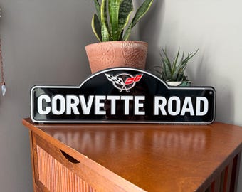 Könnte beinhalten: Schwarz-weißes Metallschild mit dem Text "Corvette Road" in weißen Buchstaben. Das Schild hat eine abgerundete Oberseite mit einem roten und weißen Corvette-Logo.