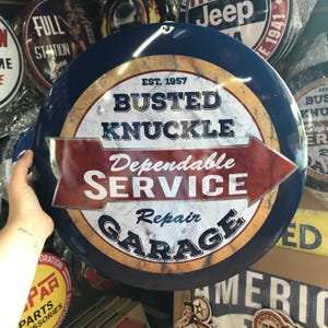 Puede incluir: Un letrero met&aacute;lico redondo y azul con el texto "BUSTED KNUCKLE Dependable SERVICE Repair GARAGE" en blanco y rojo. El letrero tiene un aspecto vintage y desgastado, con el a&ntilde;o "EST. 1957" en la parte superior. Otros letreros son visibles en el fondo.