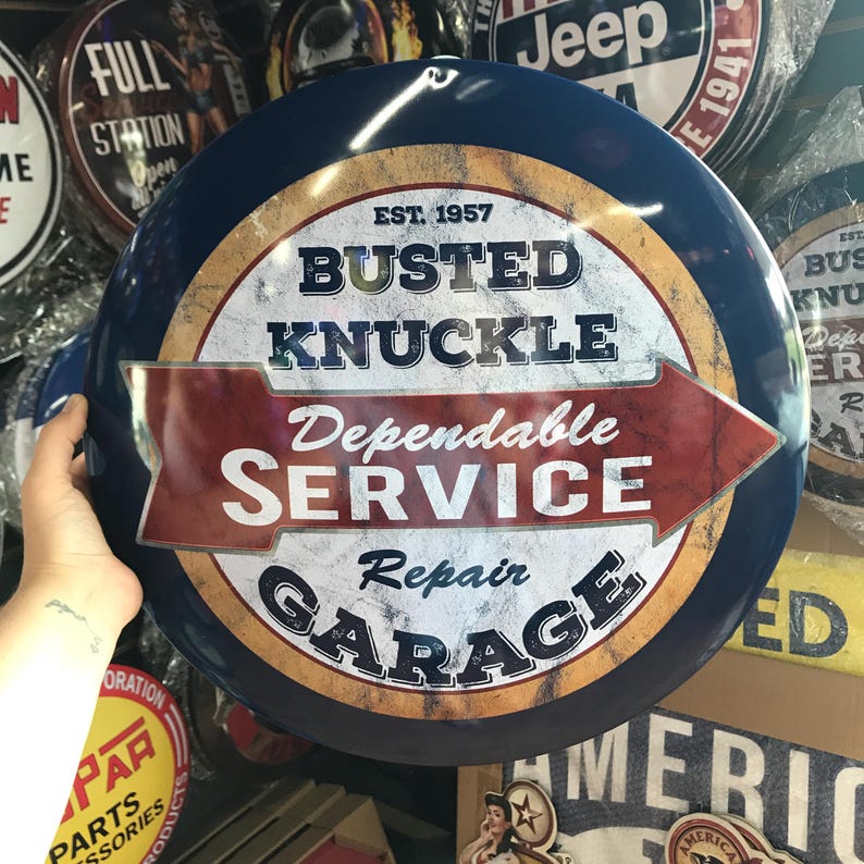 Puede incluir: Un letrero met&aacute;lico redondo y azul con el texto "BUSTED KNUCKLE Dependable SERVICE Repair GARAGE" en blanco y rojo. El letrero tiene un aspecto vintage y desgastado, con el a&ntilde;o "EST. 1957" en la parte superior. Otros letreros son visibles en el fondo.