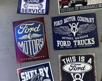 Könnte beinhalten: Eine Sammlung von Vintage-Metallschildern mit dem Ford-Logo und verschiedenen Slogans. Die Schilder sind in Blau, Rot, Weiß und Schwarz mit einem Used-Look. Die Schilder beinhalten "Genuine Parts & Ford Service", "Ford Motors", "F-Series Trucks Decades of Dominance", "Ford Motor Company 1917 2017 Detroit, Michigan Ford Trucks 100 Years of Toughness", "This is a Ford Garage" und "Shelby American".
