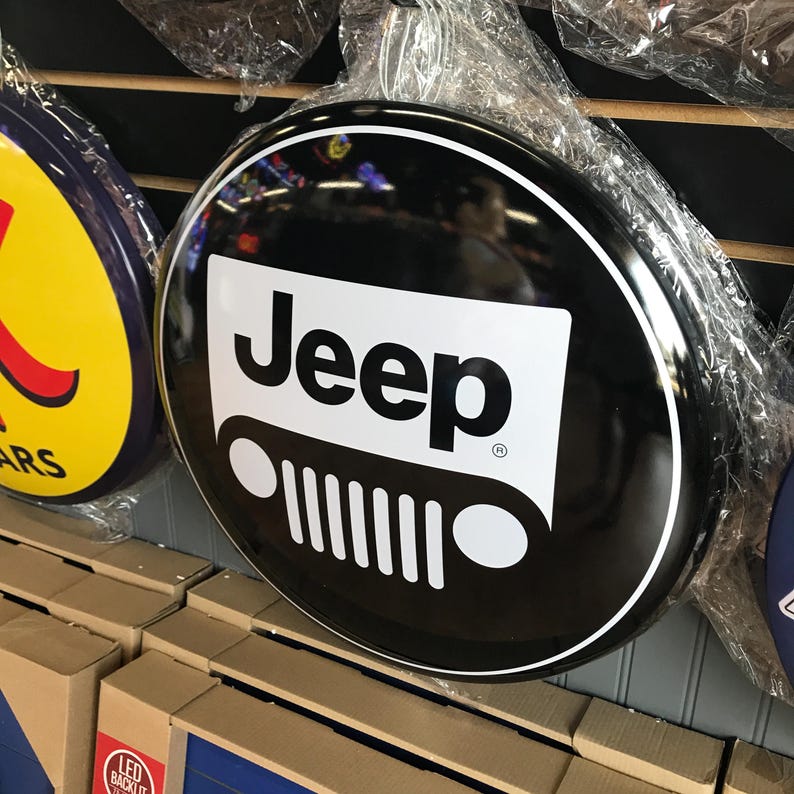 Puede incluir: Un letrero redondo de metal negro con un logotipo de Jeep en blanco y negro. El logotipo presenta la palabra "Jeep" en letras blancas y una silueta blanca de una parrilla de Jeep.