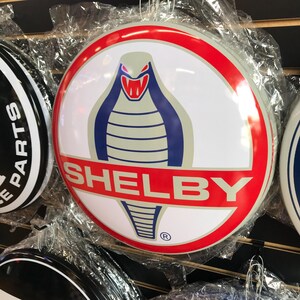 Puede incluir: Un letrero circular blanco y rojo con una cobra azul y gris con una boca roja y la palabra "SHELBY" en letras rojas. El letrero tiene un s&iacute;mbolo de marca registrada en la esquina inferior derecha.