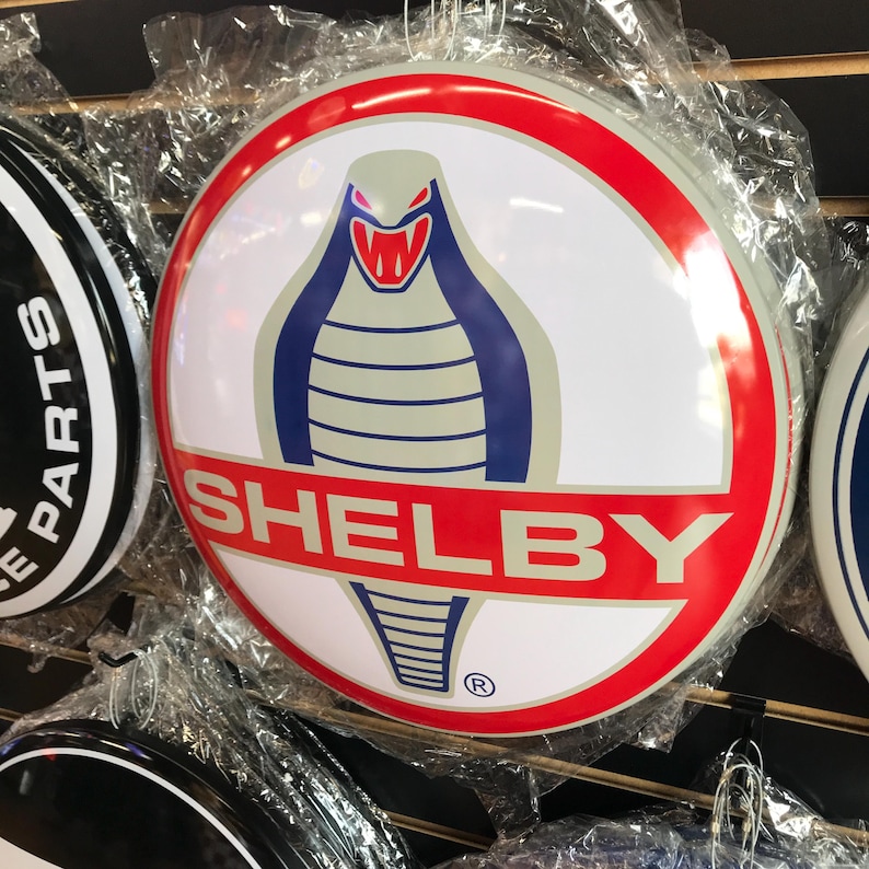 Puede incluir: Un letrero circular blanco y rojo con una cobra azul y gris con una boca roja y la palabra "SHELBY" en letras rojas. El letrero tiene un s&iacute;mbolo de marca registrada en la esquina inferior derecha.