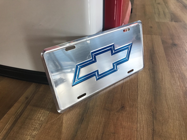 May include: A silver metal license plate with a blue and white Chevrolet logo. The plate is slightly curved and has a shiny finish.