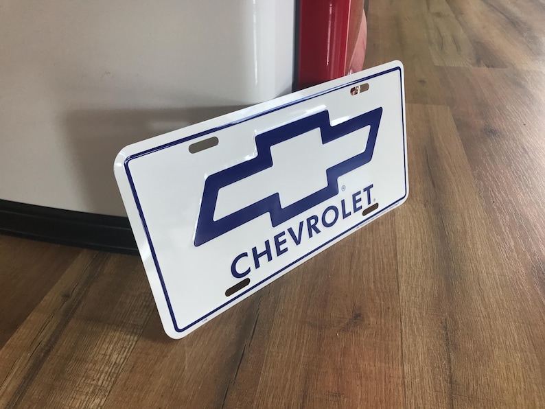 May include: White metal license plate with a blue Chevrolet logo and the word "CHEVROLET" in blue letters.