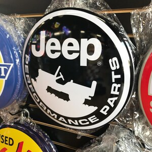 Puede incluir: Un letrero redondo de metal negro y blanco con el texto "Jeep Performance Parts" en un c&iacute;rculo alrededor de una silueta de un Jeep. El letrero tiene un fondo negro y letras blancas.