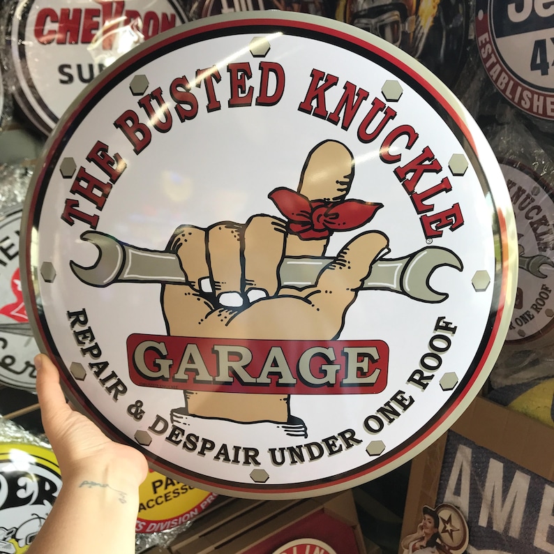 Puede incluir: Letrero met&aacute;lico redondo con el texto "THE BUSTED KNUCKLE GARAGE" en rojo y blanco. Muestra una mano de dibujos animados sosteniendo una llave inglesa, con una cinta roja en el pulgar. Con borde plateado, tem&aacute;tica de garaje.