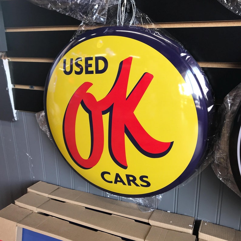 Puede incluir: Un letrero redondo de estilo vintage con un centro amarillo y un borde azul oscuro. El letrero dice "USED OK CARS" en letras rojas y negras en negrita. El letrero est&aacute; colgado y envuelto en pl&aacute;stico transparente.