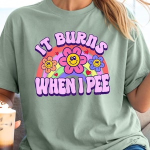 May include: A light green t-shirt with a rainbow and flower graphic. The text on the shirt reads "It burns when I pee".