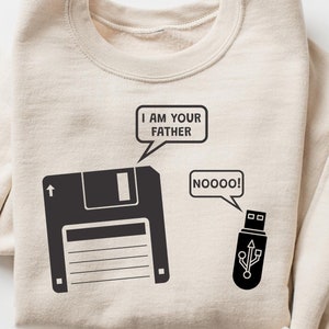 May include: Beige sweatshirt with a black graphic of a floppy disk saying "I AM YOUR FATHER" and a USB drive saying "NOOOO!". The sweatshirt has a crew neck and long sleeves. The Cozy Gifts Company logo is in the bottom right corner.