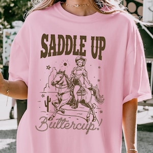 May include: Pink oversized t-shirt with a brown graphic of a woman riding a horse with the text "Saddle Up Buttercup!"