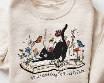 May include: Beige sweatshirt with a graphic of a black cat stretching on an open book, surrounded by flowers and butterflies. The text "It's a Good Day To Read a Book" is below the book.