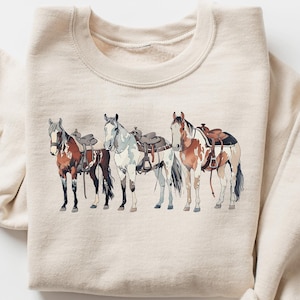 May include: A beige sweatshirt with a graphic print of four horses with saddles. The horses are brown, white, and black.
