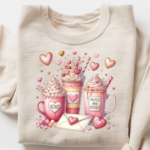 May include: Beige sweatshirt featuring a Valentine's Day design. The graphic includes three pink mugs and cups with whipped cream, hearts, and the phrases "XOXO" and "Hugs and Kisses". An envelope and hearts complete the design.