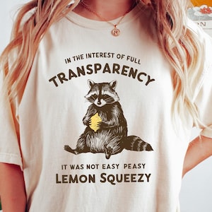 May include: A white t-shirt with a brown raccoon graphic holding a yellow lemon. The text on the shirt reads "In the interest of full transparency, it was not easy peasy lemon squeezy."