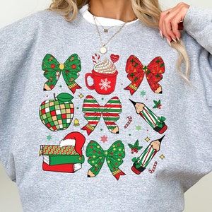 Puede incluir: Sudadera gris con un dise&ntilde;o navide&ntilde;o con lazos rojos y verdes, l&aacute;pices, una taza de chocolate caliente y una manzana.