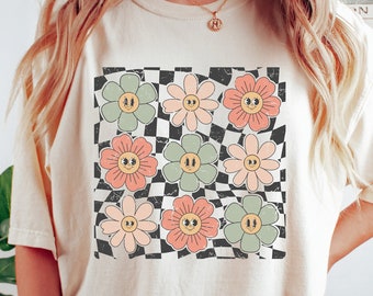May include: A cream coloured t-shirt with a checkered pattern and smiling flower graphic. The flowers are in shades of pink, green, and orange.