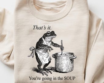 Op de afbeelding: Beige sweatshirt met een zwart-wit illustratie van een kikker met een schort die in een pot roert. De tekst "That's it. You're going in the SOUP" staat erop. Het sweatshirt is opgevouwen.