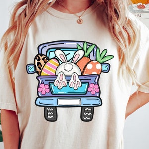 May include: Cream-colored t-shirt featuring a cartoon Easter truck filled with eggs, carrots, and a bunny. The truck is light blue with black tires and pink flowers. The bunny's white rear and paws are visible. The text "Cozy Gifts Company" is at the bottom.