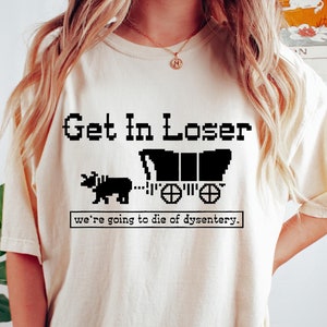 Puede incluir: Una camiseta blanca con un gráfico negro que dice "Get In Loser... we're going to die of dysentery." El gráfico es una imagen pixelada de un carro cubierto tirado por un toro.
