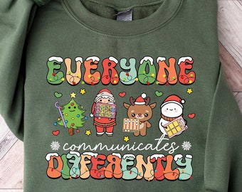 May include: A dark green sweatshirt featuring the text "Everyone communicates differently" in a retro font. The design includes festive characters like Santa, a reindeer, and a snowman, each with puzzle piece patterns.