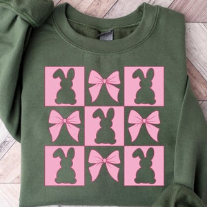 May include: An olive green sweatshirt with a pink and white checkered pattern of bunny and bow silhouettes. The design is centered on the front. The sleeves are cuffed. The bottom right corner of the image has the text "Cozy Gifts Company".