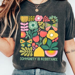Puede incluir: Una camiseta gris oscuro con un diseño floral colorido y el texto "Community is Resistance".