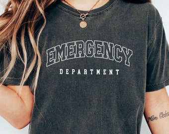 May include: A dark gray t-shirt with the text "EMERGENCY DEPARTMENT" in white outlined letters. The shirt is worn by a person with long brown hair and a gold necklace.