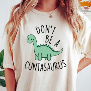 Op de afbeelding: Een wit T-shirt met een groene dinosaurus illustratie en de tekst "Don't be a Cuntasaurus".