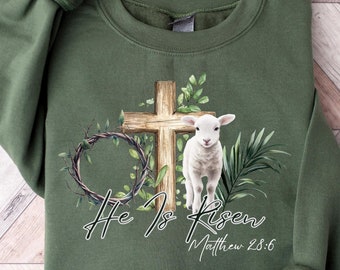 May include: Green sweatshirt featuring a white lamb standing in front of a wooden cross with a wreath of thorns and leaves. The text "He is Risen Matthew 28:6" is printed below the lamb.