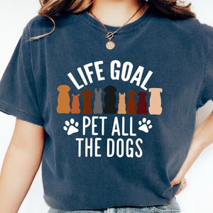 May include: A woman wearing a navy blue t-shirt with the text "Life Goal Pet All The Dogs" and a row of colorful dog silhouettes. She is wearing blue jeans and white sneakers.