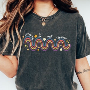 Puede incluir: Una camiseta gris oscuro con un gráfico colorido que dice "Progress is not linear" con una línea ondulada y estrellas y flores.