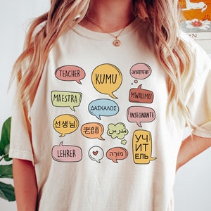 Może przedstawiać: Jasnobeżowy t-shirt z nadrukiem graficznym w postaci kolorowych dymków. Każdy dymek zawiera słowo "nauczyciel" w innym języku, w tym "Teacher", "Kumu", "Maestra", "Mwalimu", "Daskalos", "Insegnante", "선생님", "老师", "مدرس", "Уч Ит ЕЛЬ", "Lehrer" i "מוֹרָה".