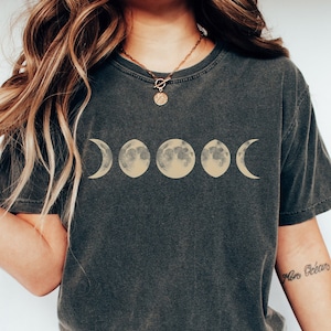 May include: A dark grey t-shirt featuring a graphic of five moon phases in shades of yellow. The shirt has a relaxed fit and a crew neck.
