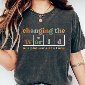 May include: A gray t-shirt with the text "changing the world one phoneme at a time" printed on it. The word "world" is in a box with each letter in a different color. There is a heart symbol between the "o" and the "r".