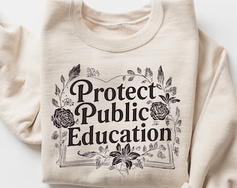 May include: Cream-colored sweatshirt with the text "Protect Public Education" in a black, floral-framed design. The long sleeves are rolled up, and the sweatshirt is folded. The design includes roses and leaves.