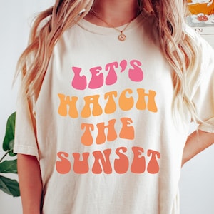 Może przedstawiać: Biała koszulka z napisem "Let's Watch The Sunset" w różowym, pomarańczowym i żółtym retro foncie.