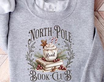 May include: Gray sweatshirt with a graphic design of a mug of hot cocoa with whipped cream and a gingerbread man, surrounded by books and greenery. The text reads "North Pole Book Club, Hot Cocoa Served Daily."