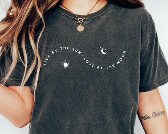 Op de afbeelding: Een donkergrijs T-shirt met een witte afbeelding waarop staat "Live by the sun. Love by the moon." De afbeelding toont een zon en een wassende maan.