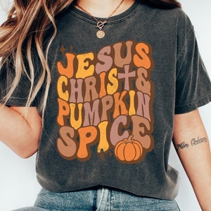 Może przedstawiać: Szary t-shirt z nadrukiem w stylu retro, na którym widnieje napis "Jesus Christ & Pumpkin Spice" w kolorowych literach. W projekcie znajduje się również mała pomarańczowa dynia.