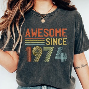 Pu&ograve; includere: Una maglietta grigia scura con il testo "Awesome Since 1974" in un design arcobaleno retr&ograve;.