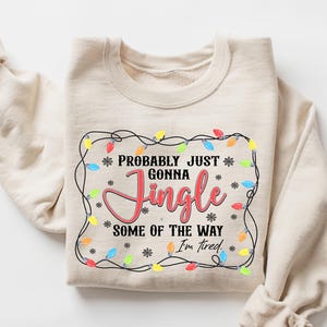 May include: A cream-coloured sweatshirt featuring the phrase "Probably Just Gonna Jingle Some of the Way I'm Tired". The design includes a border of colourful fairy lights and snowflakes, ideal for Christmas.