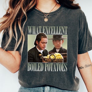 Puede incluir: Una camiseta gris oscuro con un aspecto vintage que presenta un gráfico de dos hombres con ropa de época sosteniendo un tazón de papas. El texto "What Excellent Boiled Potatoes" está impreso sobre el gráfico.