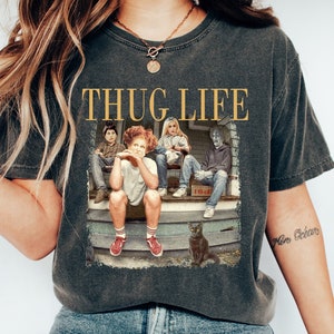 以下が含まれることがあります： 映画のキャラクターのグラフィックと金色の文字「THUG LIFE」が特徴の濃いグレーのTシャツ。 ヴィンテージスタイルで、リラックスしたフィット感です。